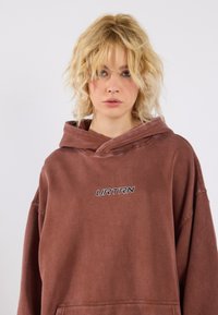 Hnedý oversized mikina s kapucňou, ktorá má prednú kapsu a výšivku s nápisom "URTRN." Mäkká tkanina so slabým opotrebovaným vzhľadom.