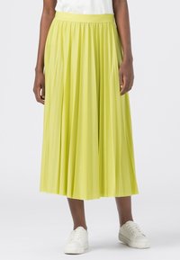 Bas du corps d'une personne portant une jupe midi plissée jaune et des baskets blanches, se tenant devant un fond clair.