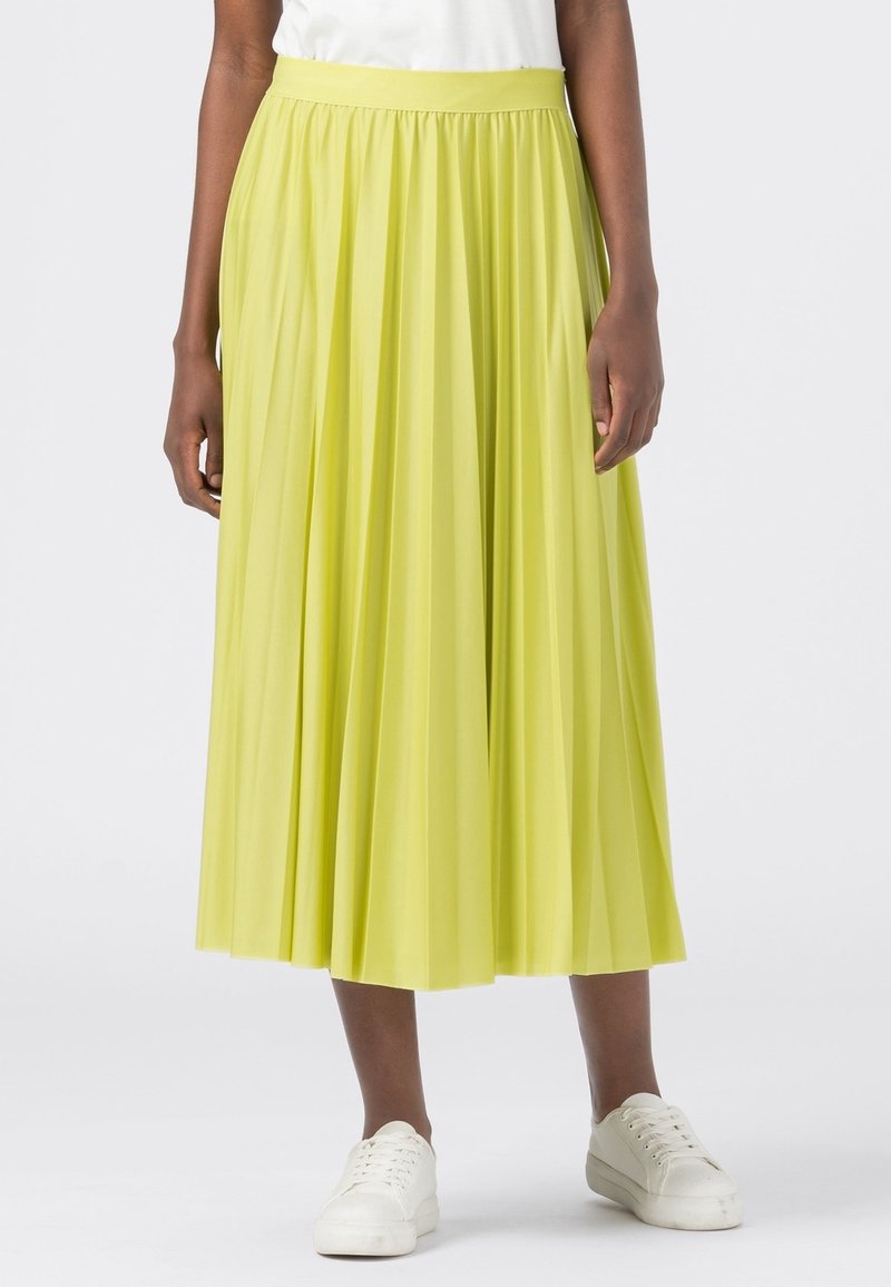 Bas du corps d'une personne portant une jupe midi plissée jaune et des baskets blanches, se tenant devant un fond clair.