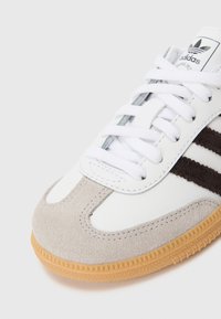 Witte sneaker met grijs suède en zwarte strepen, voorzien van een rubberen gumzool, platte veters en een merklabel op de tong. Gestructureerd en sportief ontwerp.