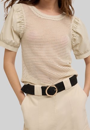 Vrouw draagt een beige top met pofmouwen onder een beige gehaakt shirt met korte mouwen, beige hoge taille broek en een zwarte riem met een ronde gouden gesp.
