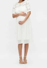 Robe de maternité blanche avec des manches courtes en dentelle et une jupe plissée. Assortie à des sandales beiges à lanières. Texture lisse, design arrivant au genou.