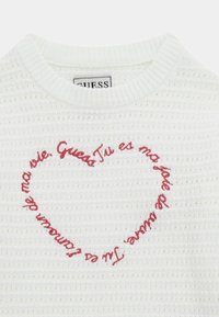 Biały sweter z teksturą, z okrągłym dekoltem i czerwonym haftowanym sercem z tekstem w środku. Widoczna metka marki przy szyi.