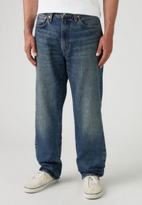 Partie inférieure du corps d'une personne portant un jean bleu ample et des baskets blanches, debout devant un fond clair uni.