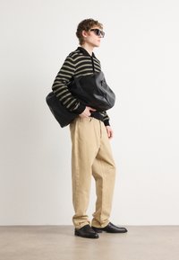 Sac de sport en cuir noir, pull rayé vert et noir, pantalon beige, chaussures noires, lunettes de soleil, se tenant contre un fond neutre.