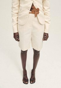 Beige maßgeschneiderte Shorts mit einem passenden Blazer, der mit einem blumenförmigen braunen Gürtel verziert ist. Einfaches Design mit Taschen. Schwarze Riemchensandalen.