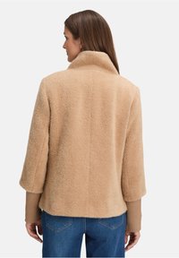 Kobieta z prostymi brązowymi włosami, w beżowej teksturowanej kurtce na jaśniejszej brązowej bluzce z długim rękawem i niebieskich dżinsach, odwrócona plecami do aparatu.