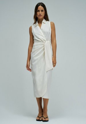 Mujer con un vestido blanco estilo wrap sin mangas, con cuello y cintura anudada, de pie, combinado con sandalias negras de punta abierta sobre un fondo liso.