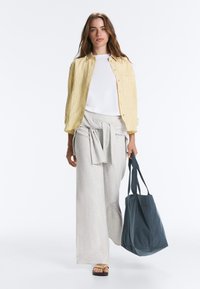 Femme portant une chemise jaune rayée sur un haut blanc, un pantalon gris ample avec un pull noué, portant un grand sac cabas bleu foncé et des sandales.