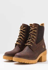 Bottines en cuir marron avec lacets jaunes, semelles épaisses et robustes de couleur jaune, talons carrés et panneaux latéraux élastiques pour un enfilage facile.