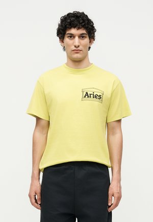 Mann mit lockigem Haar, der ein gelbes T-Shirt mit der Aufschrift "Aries" und schwarze Hose trägt, steht vor einem schlichten weißen Hintergrund.