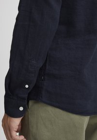 Camisa de lino de manga larga azul marino con puños abotonados y botones blancos, que presenta una etiqueta lateral. Combinada con pantalones verde oliva.