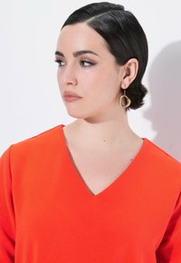 V-Ausschnitt-Oberteil aus leuchtend orangefarbenem Stoff mit strukturierter Oberfläche. Goldene, runde Ohrringe ergänzen den Look. Keine sichtbaren Muster oder Hardwaredetails.