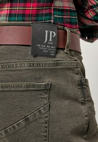 Pantaloni in denim verde con cintura in pelle marrone. Presentano dettagli cuciti, un'etichetta con "JP 1880" e dettagli sulle tasche anteriori.