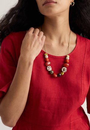 Ogrlica z biseri rdeče, zelene in bele barve z cvetličnimi vzorci, nošena prek rdečega lanenega oblačila z okroglim izrezom na vratu.
