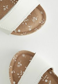 Sandales à lanières blanches avec semelles brunes ornées de motifs floraux blancs, vues de dessus sur un fond clair uni.