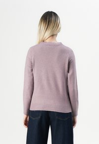 Mulher com cabelo loiro comprido vista de costas, vestindo um suéter roxo claro e calças largas azuis escuras, contra um fundo branco.
