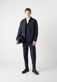 Chaqueta de puffer azul marino con dos bolsillos en el pecho, superpuesta a un suéter oscuro. Los pantalones azul marino y las botas de cuero negras hasta el tobillo completan el atuendo.