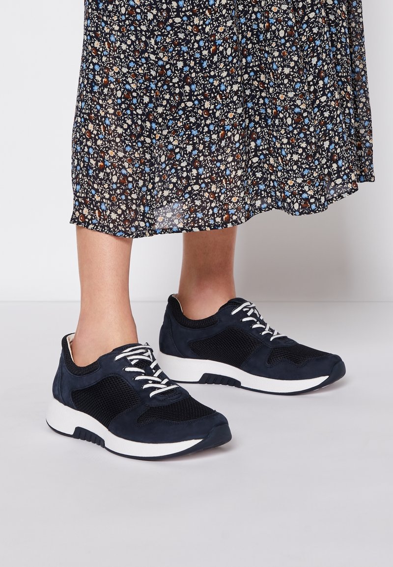 Jambes portant des sneakers en mesh noir et blanc avec des lacets blancs, accompagnées d'une jupe midi florale noire sur fond blanc.