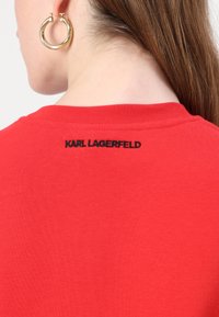 Červené bavlnené tričko s okrúhlym výstrihom. Na zadnej strane má čierny vyšívaný nápis "KARL LAGERFELD". Hladká textúra a klasický strih.