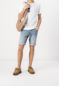 Muž v bílém tričku s potiskem, světle modrých denimových šortkách s rolovanými nohavicemi, béžových semišových botách a nese béžovou kabelku přes rameno.