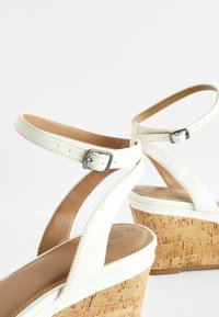 Sandales blanches à talons en liège, avec des brides croisées et des boucles ajustables à la cheville, texture en cuir lisse et doublure intérieure neutre.