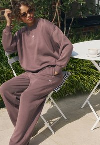 Hanorac supradimensionat mauve cu manșete din reiat, asortat cu pantaloni largi de aceeași culoare. Detaliu subtil cu logo pe hanorac și pantaloni.