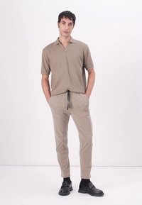 Beige polo tričko s krátkým rukávem a výstřihem do V, ke kterému nosí model volné béžové kalhoty. Model má na sobě černé boty se strapci a stojí na bílém pozadí.
