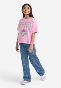 Djevojka se smije, nosi ružičasti oversized New York majicu, plave traperice s bijelim uzorcima i bijele tenisice, stoji naslonjena na bijelu pozadinu.