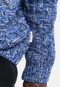 Sweter dzianinowy w niebieskich odcieniach z teksturowanym wzorem. Posiada widoczną metkę oraz ściągacze. Wykonany z grubego, przytulnego włóczki.