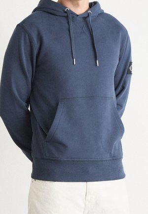 Sudadera de color azul marino hecha de tela suave, con un bolsillo frontal, cordones ajustables y puños acanalados. Incluye una etiqueta con el logo en la manga.
