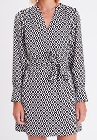 Robe à motifs noir et blanc avec un col en V, des manches longues et une ceinture à la taille. Le design présente des formes en losange et une texture de tissu fluide.