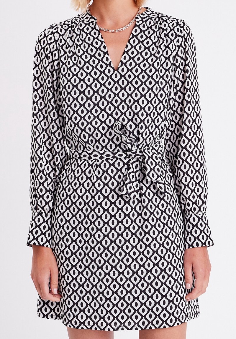 Robe à motifs noir et blanc avec un col en V, des manches longues et une ceinture à la taille. Le design présente des formes en losange et une texture de tissu fluide.