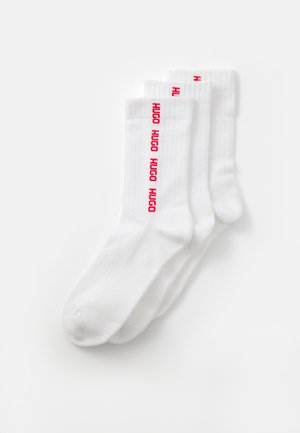 Drei Paar weiße gerippte Crew-Socken gestapelt, jeweils mit rotem "HUGO"-Text, der vertikal nahe dem oberen Bündchen verläuft.