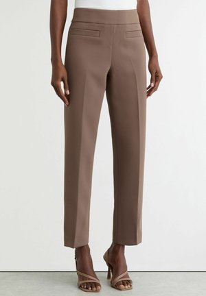 Nő magasderekú, barna, szabott nadrágot visel, elöl hajtásokkal és szűkített szárakkal, bézs pántos magassarkú szandállal párosítva.