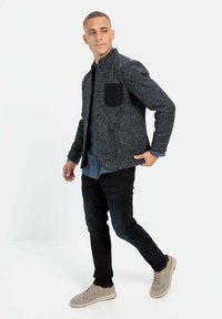 Grijze textuurjas met zwarte zak, gedragen over een blauwe shirt, in combinatie met zwarte jeans en lichtgrijze sneakers. Model staat in profiel.