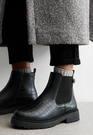 Bottines Chelsea noires en cuir à texture crocodile, avec des panneaux élastiques sur les côtés, une semelle épaisse et une tirette au dos. Portées avec des chaussettes grises à côtes.