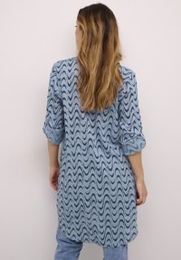 Jasnoniebieska tunika z falistym czarnym wzorem, z długimi rękawami podwiniętymi na mankietach, w luźnym kroju, wykonana z gładkiego materiału.