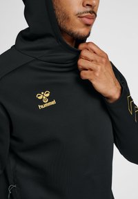 Zwarte hoodie met hoge nek en verstelbare capuchon, voorzien van een gouden "hummel"-logo op de borst en een textiel met zijritsen.