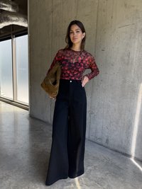 Langarmshirt mit rotem Blumenmuster, kombiniert mit hochtaillierten, schwarzen, weiten Hosen. Hält eine braune Wildledertasche. Betongrund.