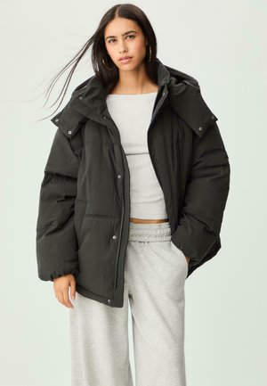 Jeune femme avec de longs cheveux noirs portant des boucles d'oreilles créoles, une grande doudoune noire, un crop top gris et un pantalon de survêtement gris sur fond uni.
