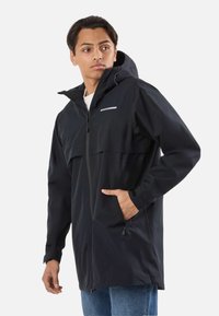 Mladý muž v čiernej nepremokavej bunde s kapucňou, so zipsom a rukou v vrecku, hľadí do boku na jednoduchom pozadí.