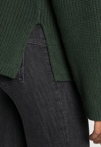 Zielony sweter o fakturowanym plecionym wzorze z bocznymi rozcięciami, noszony na ciemnych dżinsach. Materiał ma prążkowany wzór i miękki wygląd.