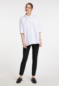 Valkoinen ylimitoitettu nappipaita lyhyillä hihoilla, yhdistettynä mustiin slim-fit-housuihin ja mustiin loafereihin, tasaisen taustan edessä.