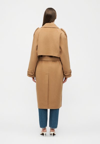 Donna in piedi di spalle che indossa un lungo cappotto color cammello con cintura, jeans blu e scarpe bianche con tacco su uno sfondo chiaro e uniforme.