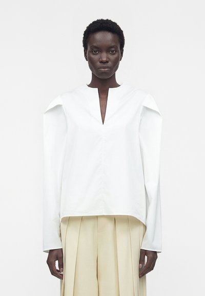 Femme aux cheveux courts et bouclés portant un haut blanc à manches longues avec des épaules rembourrées et un pantalon large plissé beige sur un fond uni.