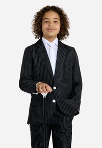 Completo a righe nere con bottoni bianchi, una camicia bianca e una cravatta argentata. La figura tiene un bastone con cappuccio bianco, in piedi con sicurezza.