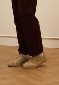 Beige suède instappers met een gestructureerde bovenkant en rubberen zolen, gecombineerd met donkerbruine broeken en witte sokken, geplaatst op een houten vloer.