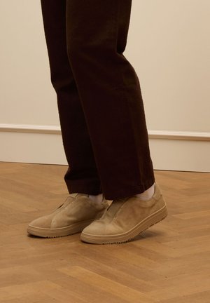 Scarpe slip-on in suede beige con tomaia texture e suole in gomma, abbinate a pantaloni marroni scuri e calzini bianchi, posizionate su un pavimento in legno.