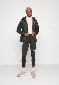 Crna vodootporna jakna, svijetloružasti gornji dio na zatvarač, tamne tajice s logom, svijetloružaste tenisice i plavi traka za glavu. Neutralna pozadina.